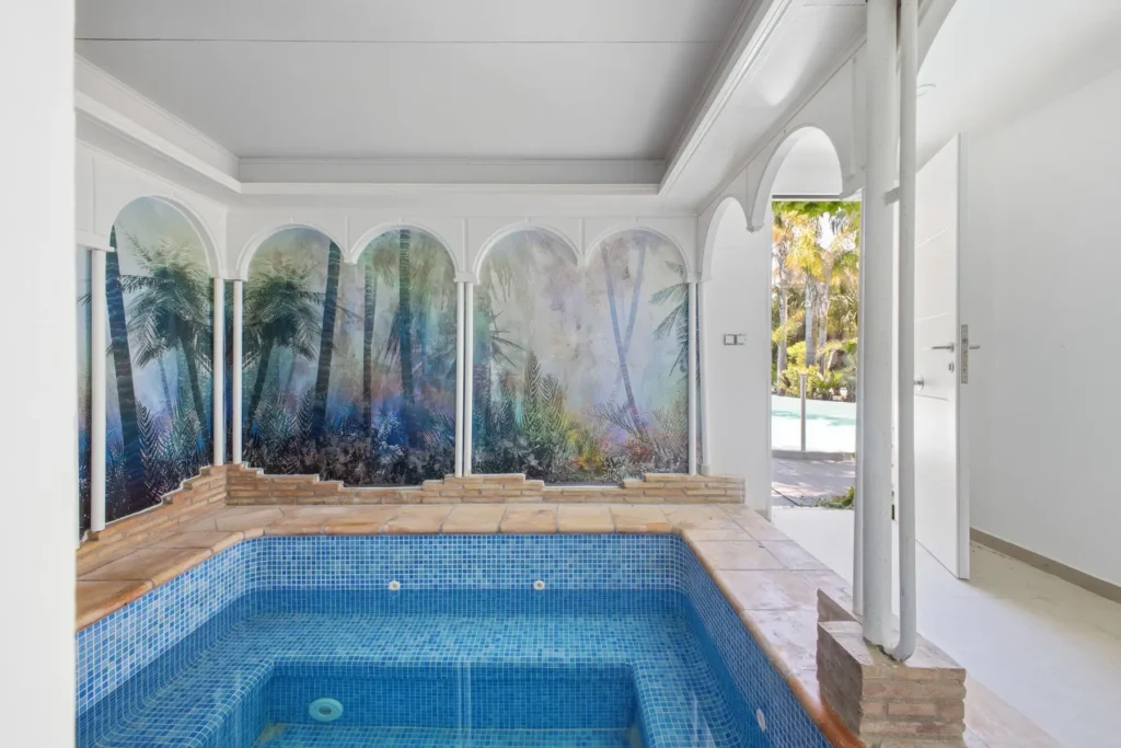 Piscina interior vacía con revestimiento azul, arcos blancos y mural de palmeras