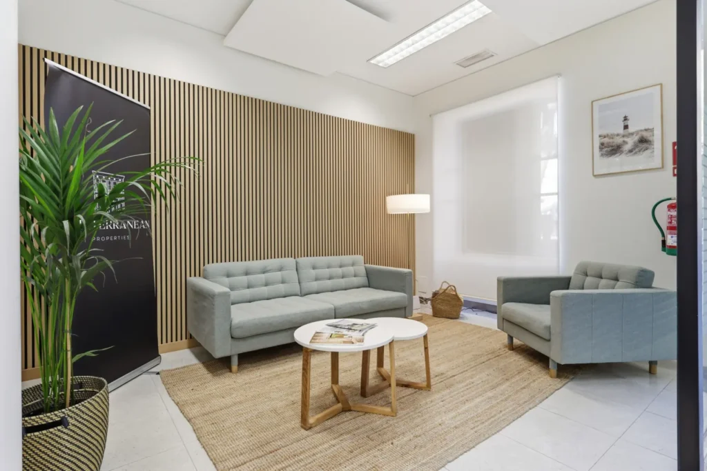Zona de recepción de una oficina con sofá gris, mesas redondas y pared de listones de madera